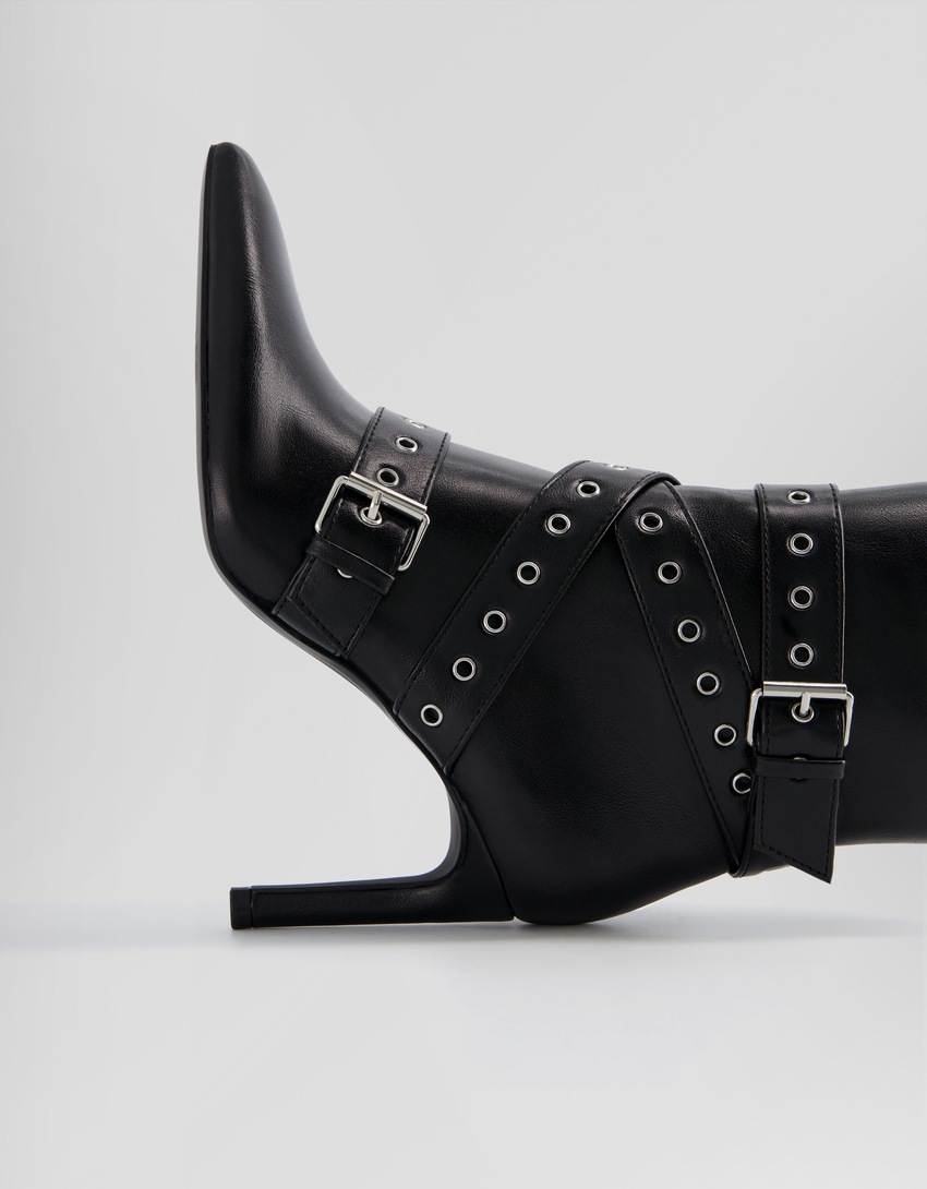 Botas de cano curto com salto agulha, tiras com ilhós e fivelas.