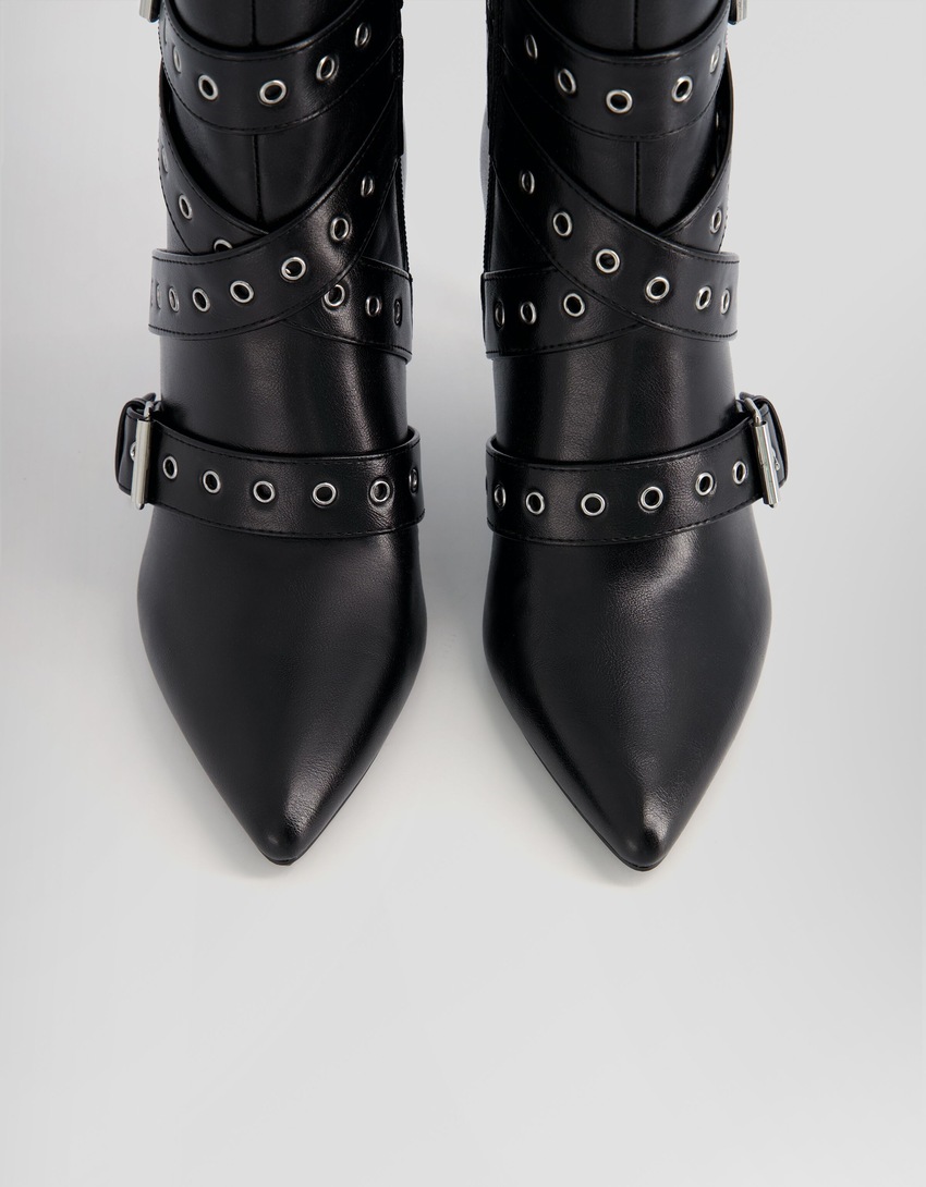 Botas de cano curto com salto agulha, tiras com ilhós e fivelas.