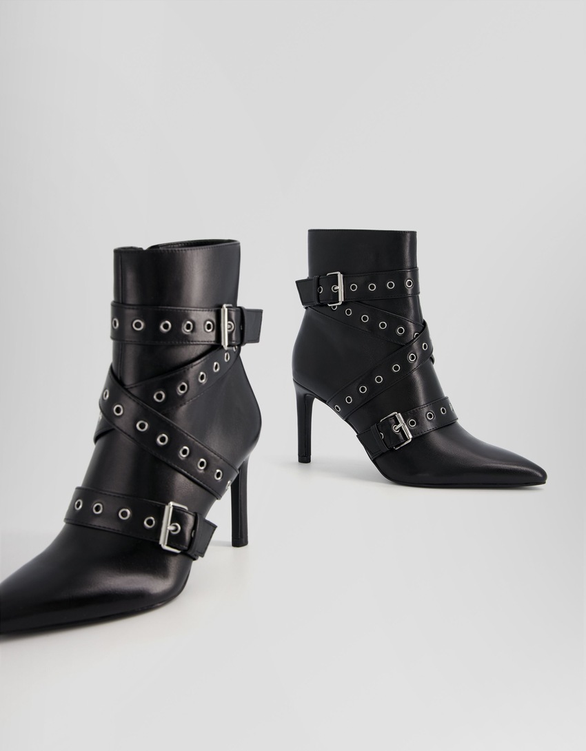 Botas de cano curto com salto agulha, tiras com ilhós e fivelas.