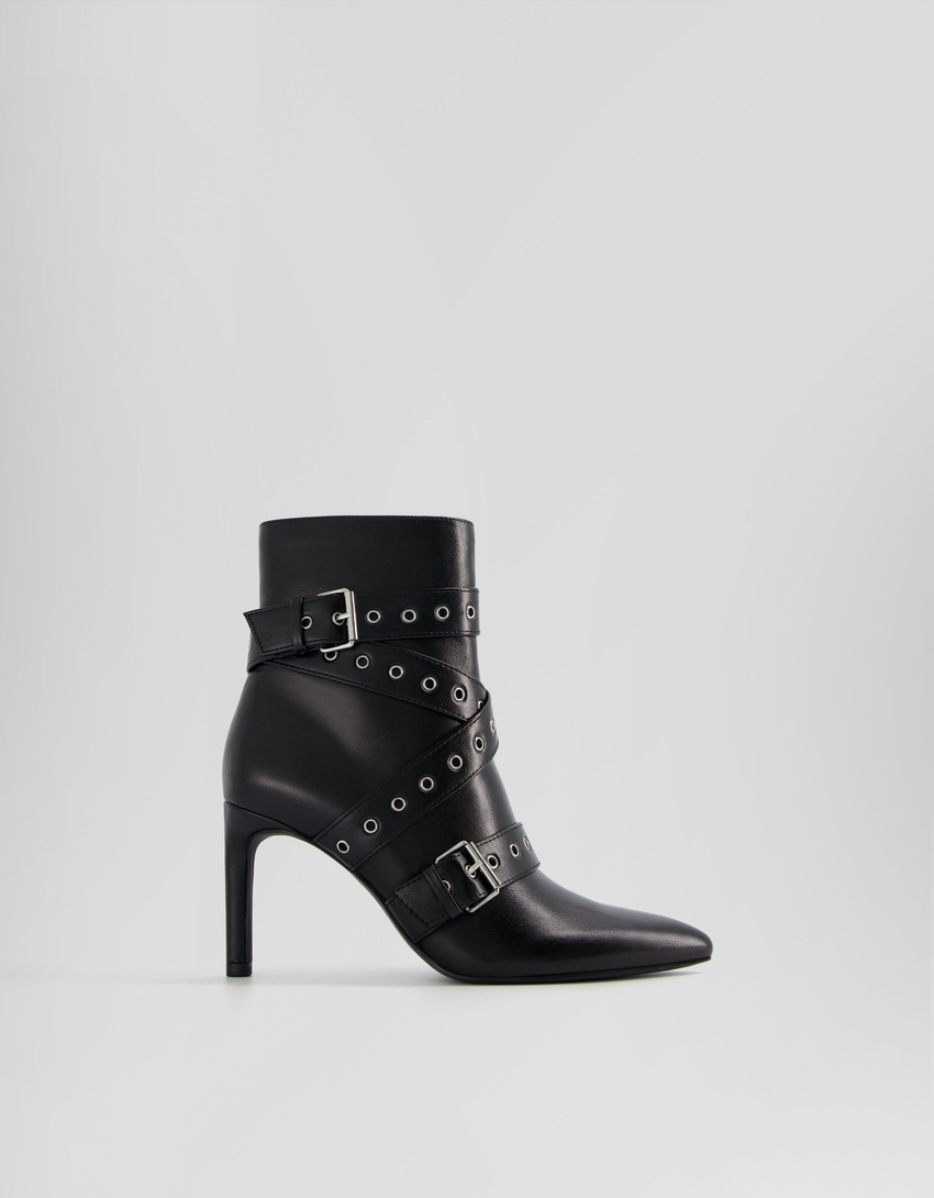 Botas de cano curto com salto agulha, tiras com ilhós e fivelas.