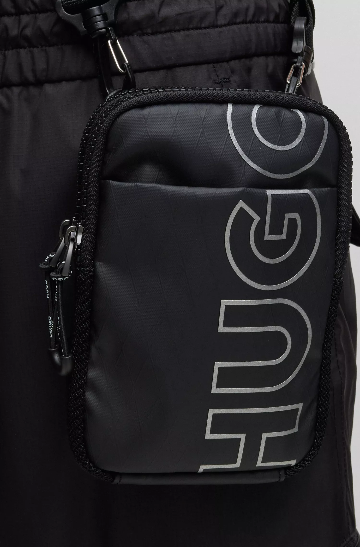 Bolsa estilo repórter HUGO com logo em contorno vermelho, confeccionada em material com textura de diamante.
