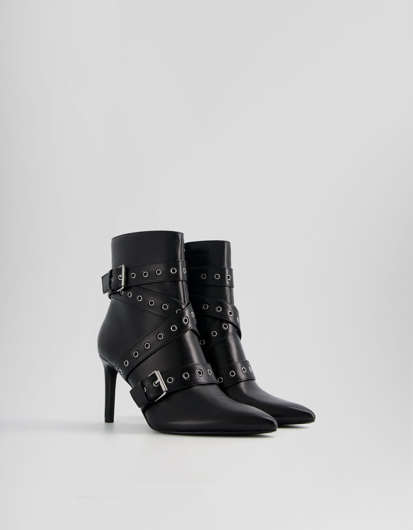 Botas de cano curto com salto agulha, tiras com ilhós e fivelas.